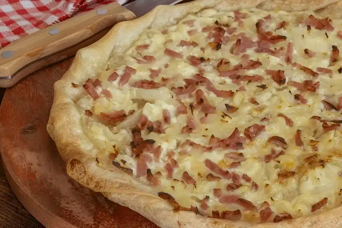 Une délicieuse tarte flambée alsacienne, garnie de crème fraîche, d'oignons et de lardons fumés, présentée sur une planche en bois.