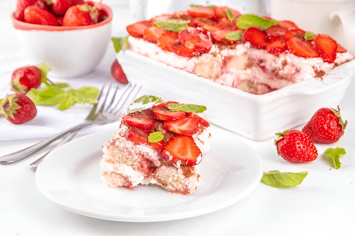 Part de tiramisu aux fraises servie sur une assiette, avec le plat complet en arrière-plan