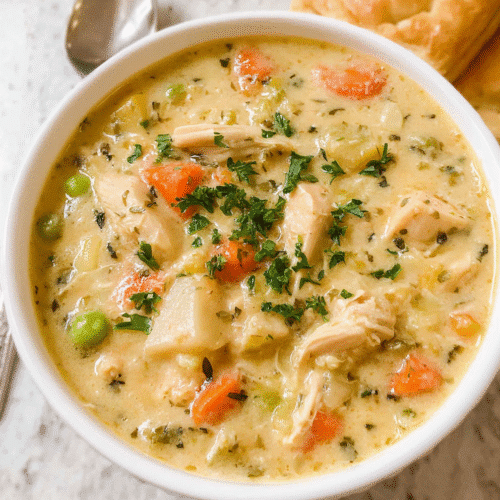 Creamy chicken pot pie soup in a white bowl, filled with carrots, peas, potatoes, and shredded chicken, garnished with fresh parsley and served with puff pastry on the side.