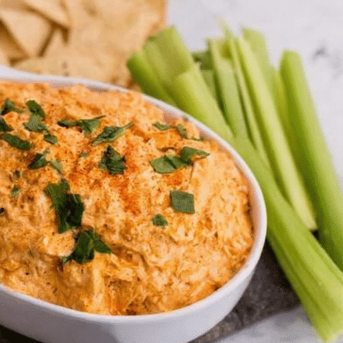 Spicy creamy buffalo chicken dip served in a white bowl, topped with herbs, with celery sticks and tortilla chips on the side, delicious party appetizer