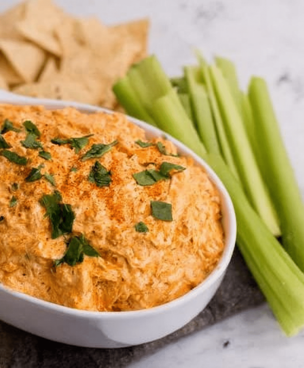 Spicy creamy buffalo chicken dip served in a white bowl, topped with herbs, with celery sticks and tortilla chips on the side, delicious party appetizer