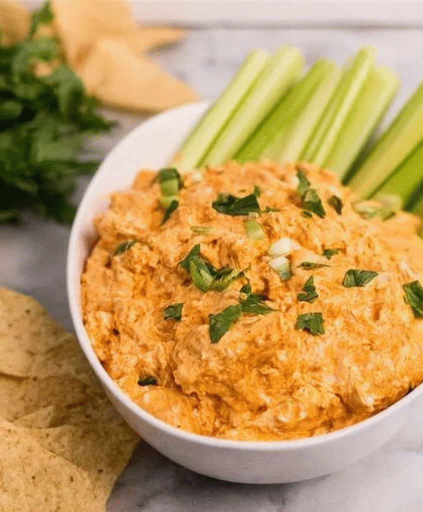 Spicy creamy buffalo chicken dip served in a white bowl, topped with herbs, with celery sticks and tortilla chips on the side, delicious party appetizer
