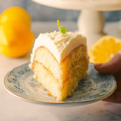 Slice of moist lemon layer cake with creamy frosting on a white cake stand in natural sunlight