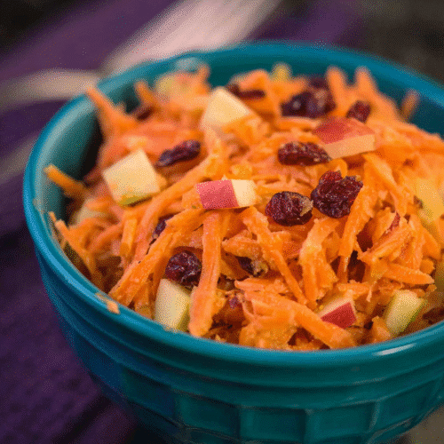Fresh shredded carrot and apple salad mixed with dried cranberries in a teal bowl on dark background, healthy and vibrant.