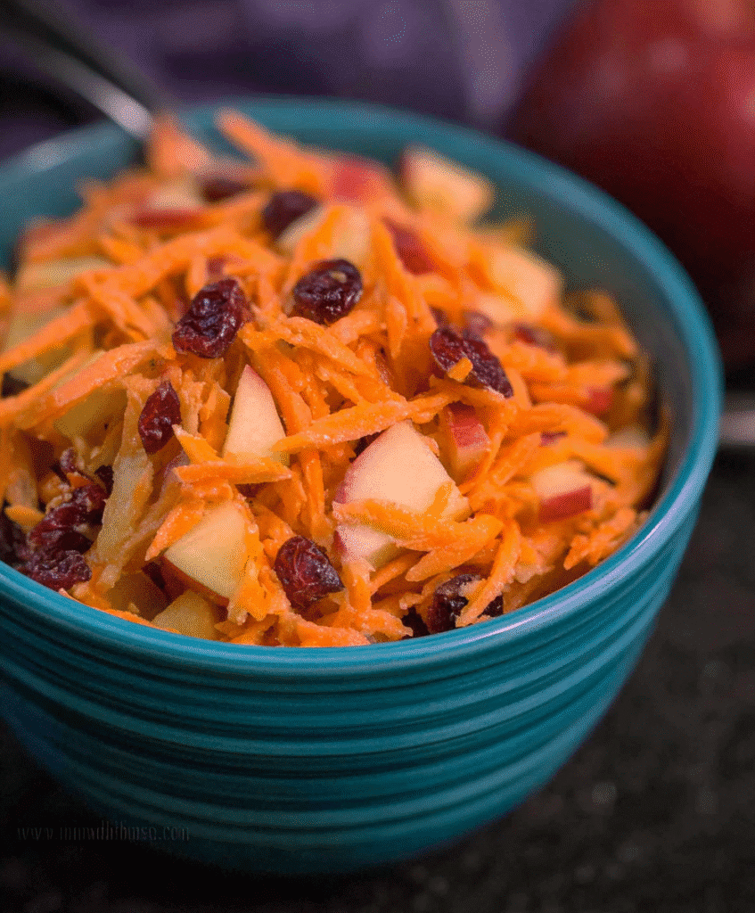 Fresh shredded carrot and apple salad mixed with dried cranberries in a teal bowl on dark background, healthy and vibrant