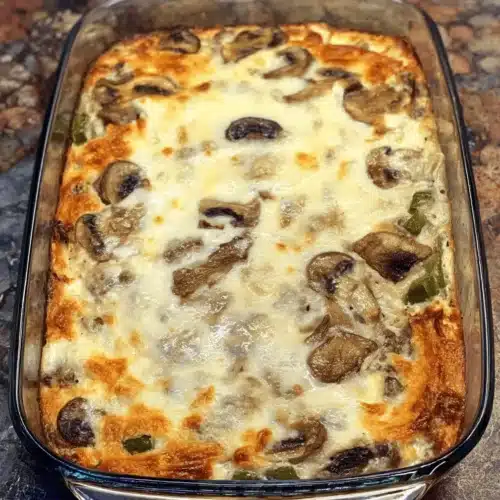 Freshly baked Low Carb Philly Cheesesteak Casserole in a glass dish, featuring a golden-brown cheesy crust, mushrooms, and green bell peppers.
