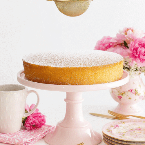 Fluffy French butter cake dusted with powdered sugar on a pink cake stand, with a hand holding a gold sifter and pink floral table decor