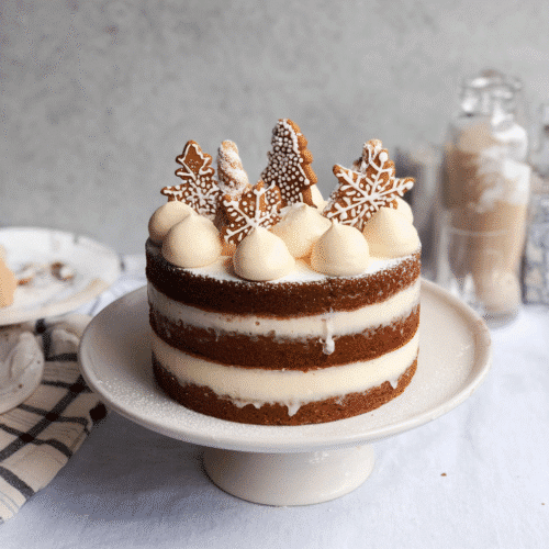 Festive gingerbread layer cake with cream cheese frosting, decorated with iced gingerbread cookies and snowy winter details on a cake stand.”